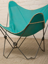 Load image into Gallery viewer, A turquoise butterfly-style chair with a black metal frame, positioned on a beige textured rug against a white brick wall.
