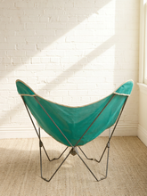 Load image into Gallery viewer, A teal butterfly chair with a black metal frame, set against a white brick wall with diagonal sunlight shadows and a beige woven rug.

