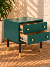 Load image into Gallery viewer, Image 4 of 5: A modern two-drawer side table in dark teal green with gold drawer knobs, black legs, and gold-toned metal foot caps, shown with two drawers partially open. The nightstand is set on a woven jute rug against a light brick wall with a potted plant in the background.
