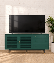 Load image into Gallery viewer, Image 2 of 5: A dark green TV stand cabinet with gold hardware, shown holding a black flat-screen TV in a styled room with a white brick wall and a potted plant.
