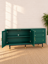 Load image into Gallery viewer, Image 4 of 5: A modern sideboard cabinet in deep teal green, shown with its left door open to reveal an interior shelf. The cabinet features three drawers on the right with brass knobs and stands on black legs with gold caps.
