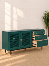 Load image into Gallery viewer, Image 5 of 5: A modern sideboard cabinet in deep teal green, featuring two louvered doors, three open drawers on the right with light wood interiors, and gold-tone hardware, set against a white brick wall with a potted plant.
