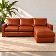 Load image into Gallery viewer, Image 1 of 14: Simulated Hollis 103-inch two-piece sectional in Mont Blanc Caramel leather with an integrated right chaise, styled on a plain ivory rug beside a bird-of-paradise plant against white brick.
