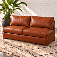 Load image into Gallery viewer, Image 10 of 14: Simulated Hollis 61-inch armless condo sofa in Mont Blanc Caramel leather with two seat cushions and two back cushions, styled with an ivory rug and bird-of-paradise plant.
