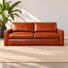 Load image into Gallery viewer, Image 2 of 3: Simulated Hollis 84-inch sofa in Mont Blanc Caramel leather with two seat cushions and two back cushions, styled on a plain ivory rug beside a bird-of-paradise plant against white brick.
